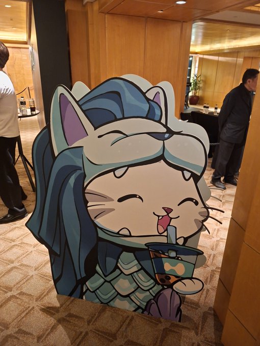 A photo of a cardboard cutout depicting a chibi cat-like character in armor drinking bubble tea, displayed in what appears to be a hotel lobby.