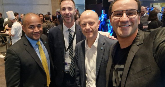 Group selfie of four men in business attire at what appears to be a professional conference, with people mingling in the background.