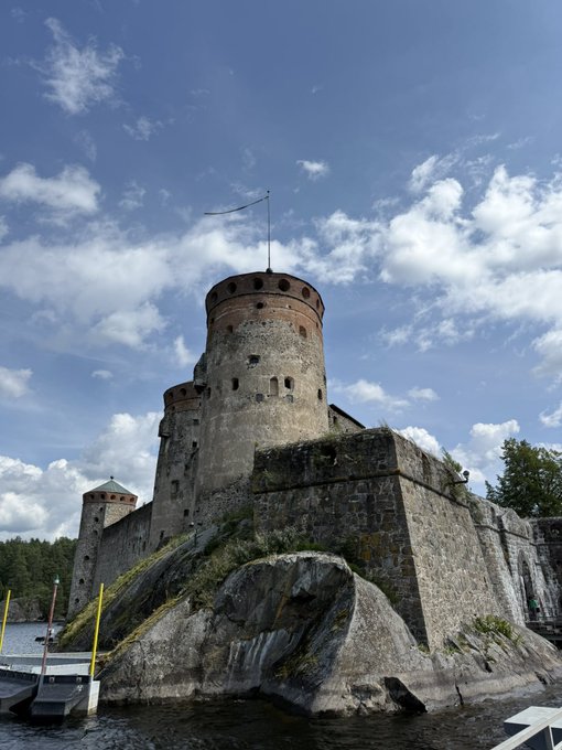 A photograph of a medieval stone fortress with cylindrical towers situated on a rocky outcrop beside a body of water under a partly cloudy sky, resembling Olavinlinna castle.