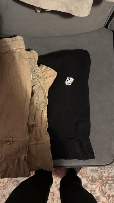 A top-down view of folded clothing on a gray surface, including a tan garment and a black hoodie with a small white logo, with a person's feet in black socks visible at the bottom.