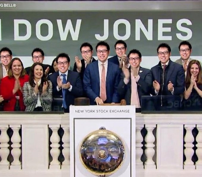 A group of people in business attire applauding at the New York Stock Exchange in front of a 'DOW JONES' sign and the ceremonial bell.