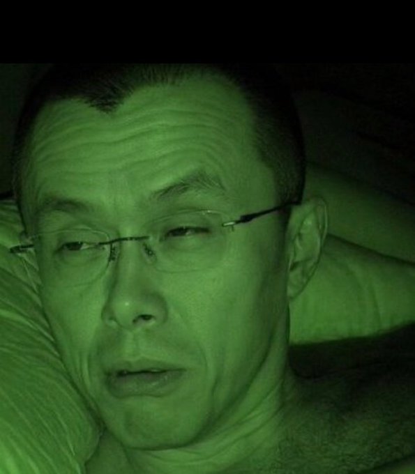 A man with glasses lies in bed, illuminated by a green night-vision hue, appearing tired or unwell. The caption's reference to a single memecoin trade altering one's life forever is humorously reflected in the man's exhausted, defeated expression—suggesting the toll of risky crypto speculation.