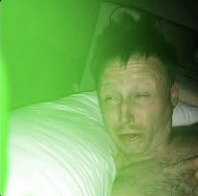 A grainy, green-tinted photo of a man with dark hair lying down against a white pillow, appearing unwell.