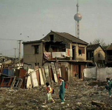 Two children stand in a debris-filled area with makeshift houses and a tall TV tower visible in the background, depicting urban poverty.