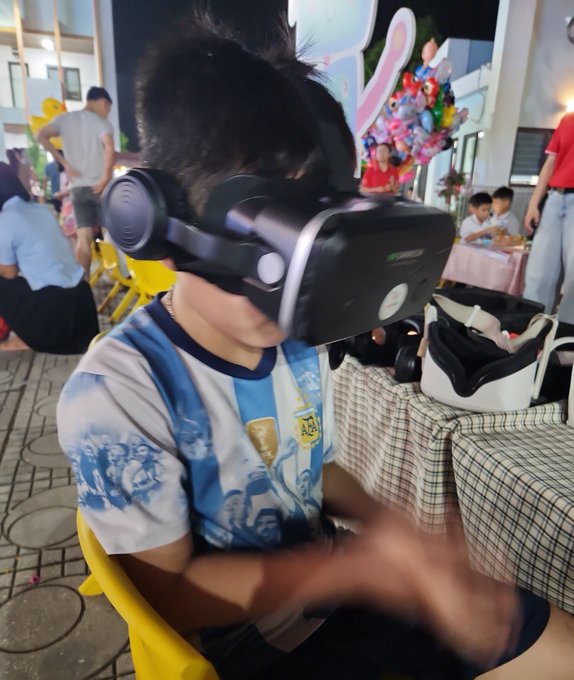 A young boy wearing a VR headset and an Argentina soccer jersey (number 43) seated at what appears to be an outdoor event with balloons in the background.