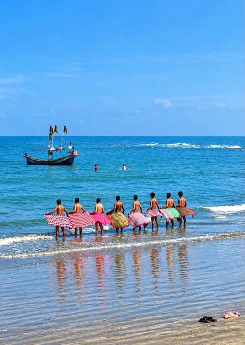 A beach scene with people carrying surfboards into the ocean while a small fishing boat floats in the background.