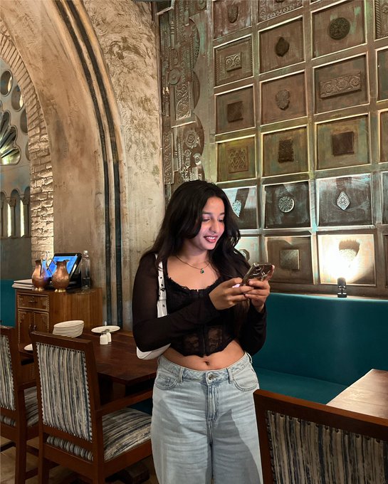 A young woman smiles down at her phone in a restaurant with ornate Middle Eastern-inspired décor featuring textured wall panels and arched doorways. The caption playfully suggests the phone displays a notification from a liquidation sale email, humorously implying she's found her next shopping target.

No readable text, numbers, tickers, or names are visible on her phone screen in the image.