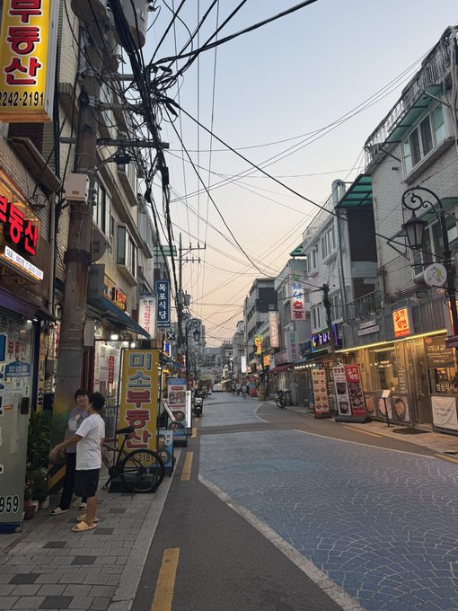 A Korean street scene at dusk with various shop signs in Korean (including one for '신호산' with 242-2191), tangled overhead power lines, and a couple of pedestrians on the sidewalk.