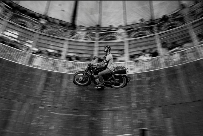 A motorcyclist wearing a helmet rides along the curved wall of a wooden "Wall of Death" arena in a black-and-white photograph, with the safety fence enclosing the daring performance. The image captures the essence of the post's theme—where the fence transforms from a mere barrier into the very structure that makes the dangerous stunt possible, embodying the boundary between dreams and dread.