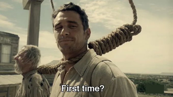 The image shows a man wearing a shirt with the words "First time?" written on it, standing next to a rope that is attached to another person's neck. The scene appears to be set outdoors or in an open area, possibly near a building.