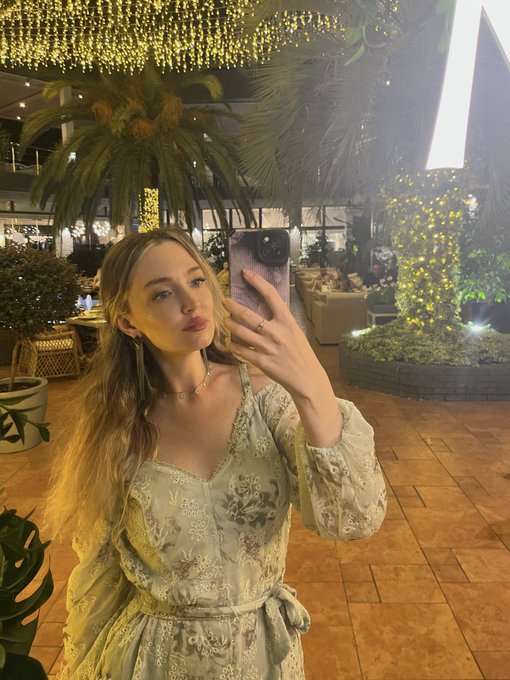 A young woman with long blonde hair takes a mirror selfie with her phone in an elegant light green floral dress, set in a warmly lit venue with hanging lights and palm trees.