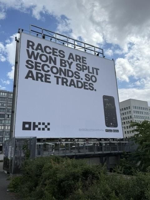 An OKX billboard stating 'RACES ARE WON BY SPLIT SECONDS. SO ARE TRADES.' displayed on a building with a partly cloudy sky.