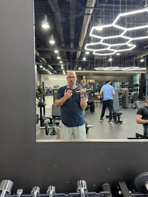 A man with a beard, glasses, and a dark t-shirt takes a selfie in a gym mirror, holding a phone and a white container, with weight equipment and another person in a blue shirt visible in the background.