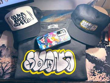 A flat lay photo showing merchandise including two caps, a t-shirt with yellow graffiti-style text, and a phone case with colorful artwork.