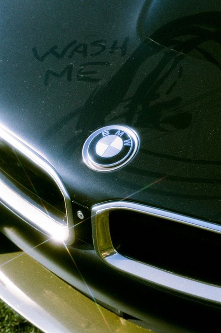 A dusty black BMW hood displays the handwritten message "WASH ME" above the iconic blue and white logo, with the context suggesting the owner received a 1/1 NFT titled "BEAMER" referencing their beloved car.