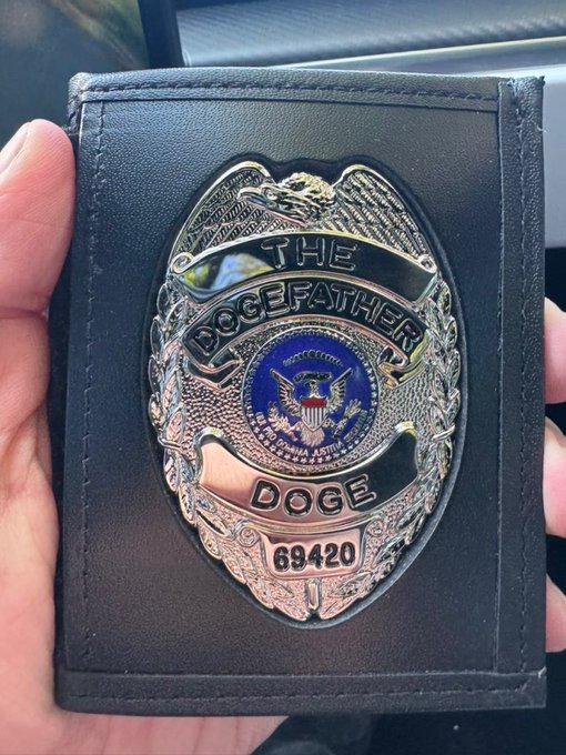The image features a person holding up an item that is shaped like a badge with the words "DOGE" written across its center, along with the phrase "THE DOGE FATHER". This badge appears to be a police or security badge. The person holds it in their hand, showcasing the details of this distinctive item.