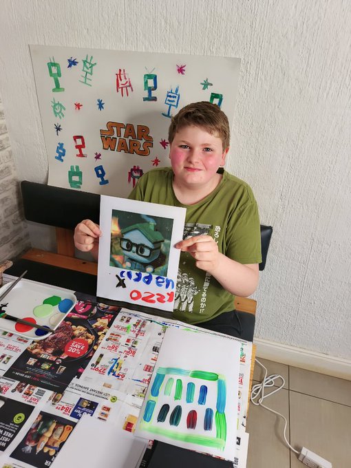 A young boy in a green Star Wars t-shirt proudly displays his artwork, including a painting of a Splatoon character and a watercolor color swatch sheet, surrounded by art supplies and a homemade Star Wars poster on the wall behind him.
