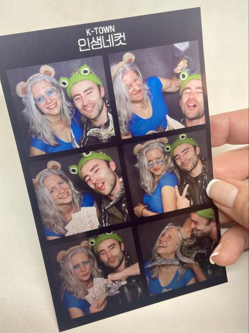 A photo booth strip from 'K-TOWN 인생네컷' featuring six pictures of a couple wearing frog-themed headbands, with the woman in a blue top and the man holding money.