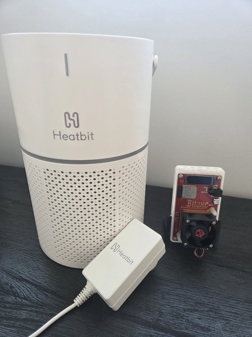 The image shows a white Heatbit-branded cylindrical device (likely the Bitair) on a dark surface, accompanied by a smaller white Heatbit power adapter and a small red Bitaxe circuit board for size comparison. The visible "Heatbit" logos on both the larger unit and the adapter match the post's description of the Bitair being shown alongside a Bitaxe reference unit at Solo Satoshi Headquarters, illustrating how compact the Bitair actually is.