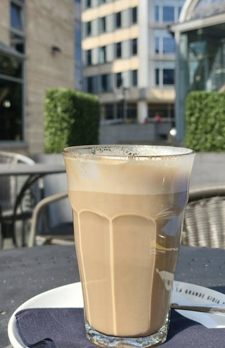 A tall glass of iced coffee with a frothy top sits on a saucer at an outdoor café table, with a blurred city building in the background on a sunny day.