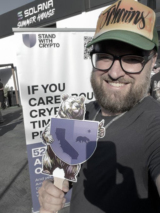 A bearded man wearing glasses and a "Tuggins" cap smiles while holding a California bear shield cutout in front of a "Solana Summer House" banner featuring "Stand With Crypto" text and a QR code. The image captures his enthusiasm at the crypto event, aligning with his post about building a project on $SOL and appreciating the Solana community gathering.