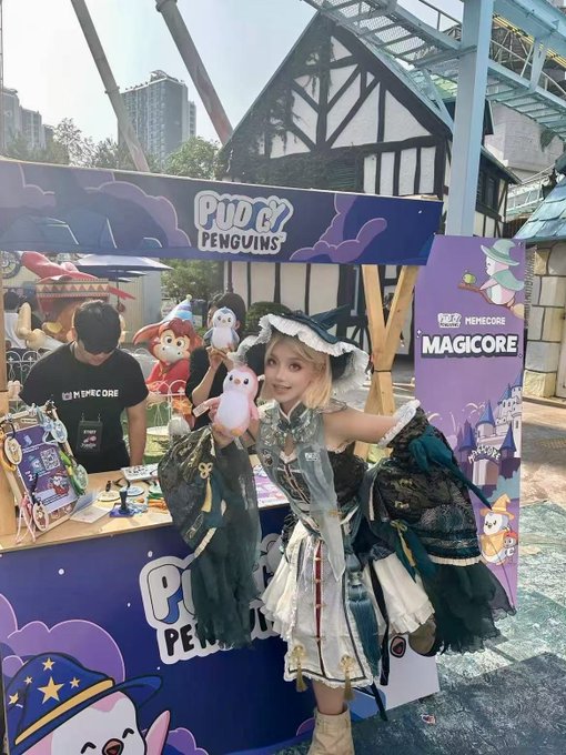 A cosplayer holding a penguin plushie at an outdoor promotional booth branded with "PUDGY PENGUINS" and "PUDGY MEMECOIN MAGICCORE" logos.