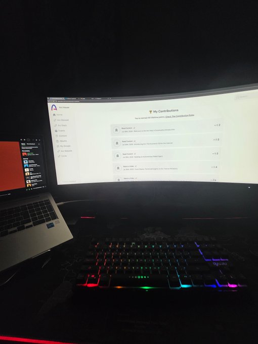 A dimly lit desk setup featuring a laptop on the left next to a large ultrawide curved monitor displaying an 'Arch Linux' page titled 'My Contributions', with an RGB mechanical keyboard in the foreground.