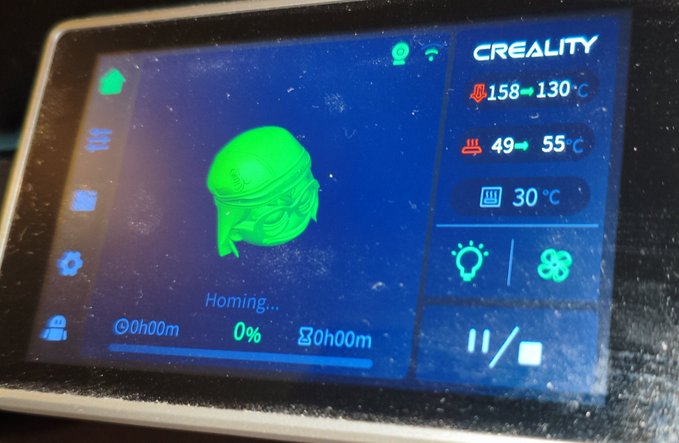 A close-up of a Creality 3D printer's touchscreen interface displaying a green ninja mask model in 'Homing...' status at 0% progress, with nozzle temperature at 158→130°C.