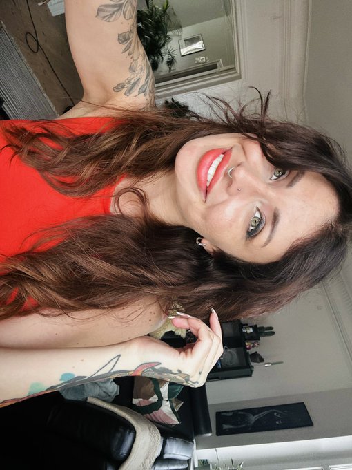 A vertical selfie of a smiling woman with long brown hair wearing a red top, with visible tattoos on her arm, taken in what appears to be a salon or studio setting.