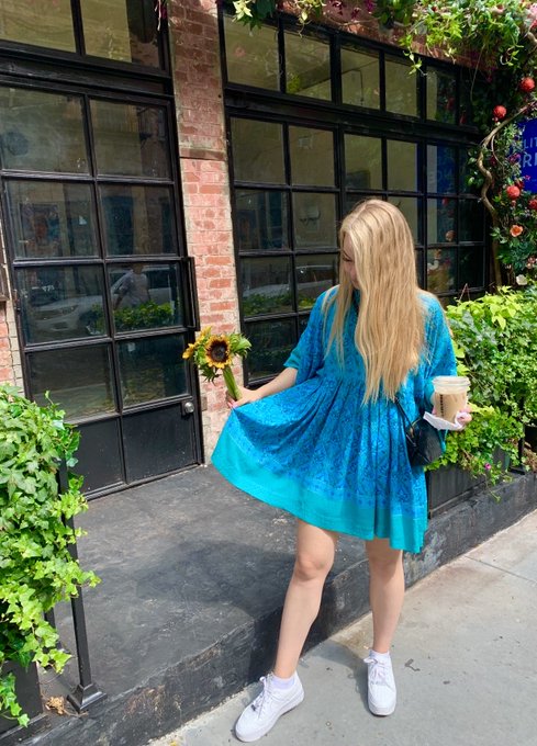 A woman in a turquoise dress holding sunflowers and a coffee cup, standing on a sidewalk in front of a brick building with large windows.