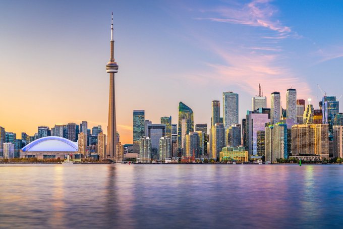 A panoramic view of the Toronto skyline at sunset, featuring the iconic CN Tower and the Rogers Centre dome along the waterfront of Lake Ontario.