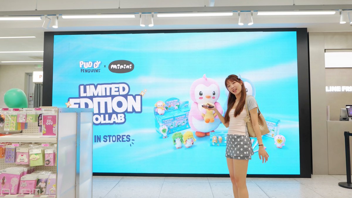 A young woman poses in front of a large illuminated billboard advertising a "Pudgy Penguins x miniso" "LIMITED EDITION COLLAB IN STORES" inside a retail store, showcasing the brand's expansion into Asian markets like the one mentioned in the caption.