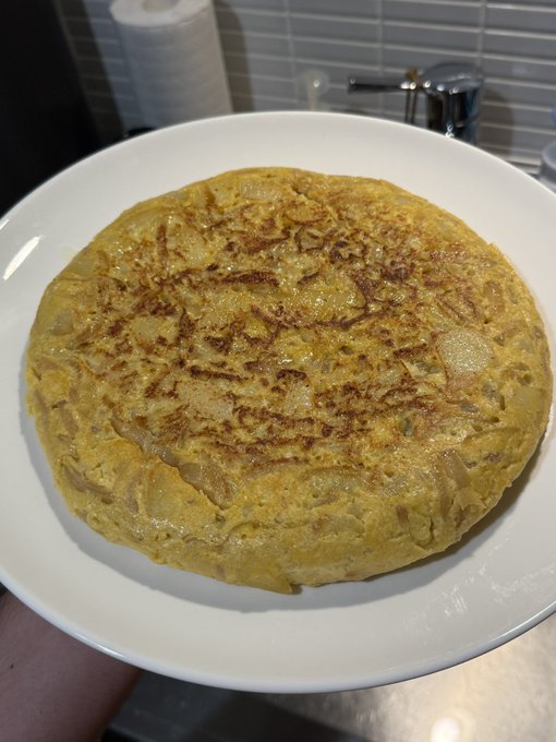 A thick Spanish tortilla (potato omelette) with melted cheese and onions sits on a white plate in a kitchen setting.