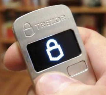 A silver Trezor hardware cryptocurrency wallet held in a hand, displaying an unlocked padlock icon on its small screen.