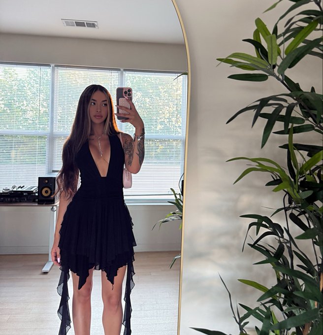 A young woman with long dark hair, tattoos, and a black dress takes a mirror selfie in a bright room with a large arched mirror and a plant nearby, with the post's "degen" caption playfully contrasting her polished, put-together appearance with a self-deprecating joke about her lifestyle.