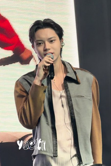 A young male performer with dark hair, wearing a white top and a grey and brown jacket, sings into a microphone on stage.