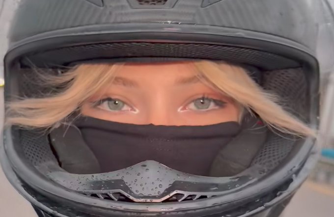 A close-up selfie of a person with blonde hair, light eyes, and makeup wearing a black face mask and a dark motorcycle helmet, with only their eyes visible.