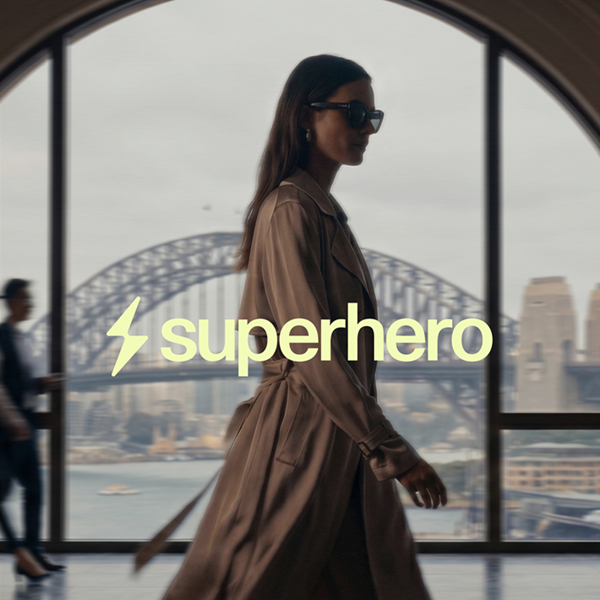 A fashion-forward image of a woman in a tan trench coat and sunglasses with the bold yellow "⚡superhero" logo overlaid, with Sydney's Harbour Bridge visible through arched windows in the background.