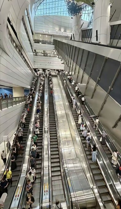 A high-angle view of a busy multi-level transit station with three long parallel escalators filled with commuters.