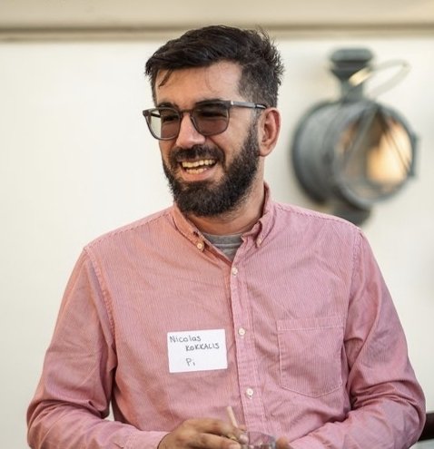 A smiling, bearded man wearing glasses and a name tag reading "Nicolas Kokkalis, Pi" stands in front of a wall with a decorative lamp, visually corresponding to the article about Nicolas Kokkalis and the PiRC1 experiment.
