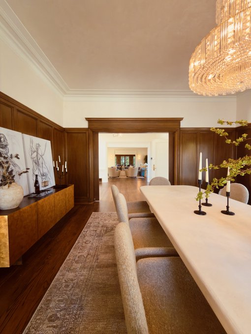An elegant dining room featuring a long table, upholstered chairs, a crystal chandelier, and wood-paneled walls, with framed sketches visible on the left.