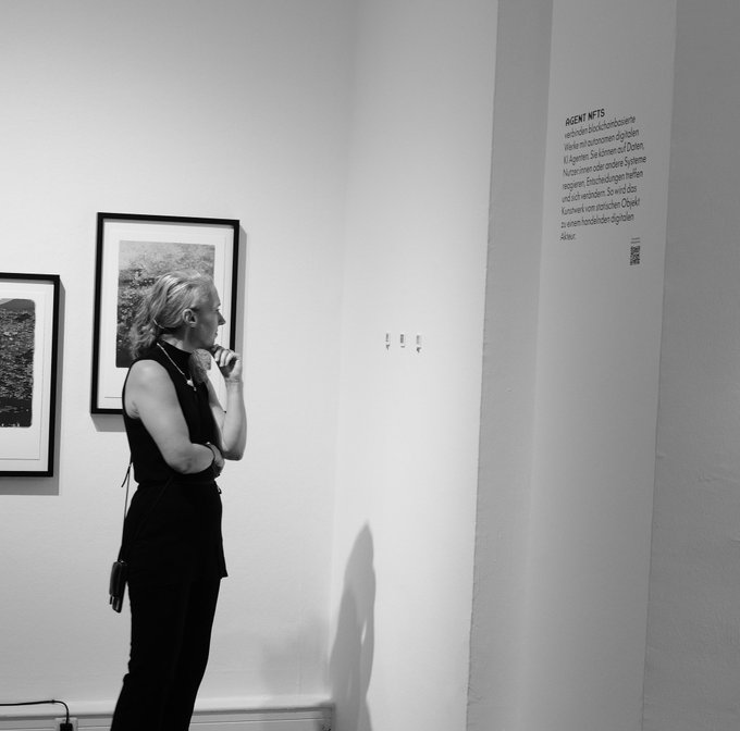 A woman with blonde hair and glasses stands in a black dress, thoughtfully viewing framed photographs in a white-walled gallery. On the right wall, text introduces "AGENT NFTS," noting that works will be auctioned on the blockchain with proceeds going to support artists and Ukraine, curated by Tschuuuuly. The image reflects the gallery's atmosphere during the large-scale NFT exhibition mentioned in the post.