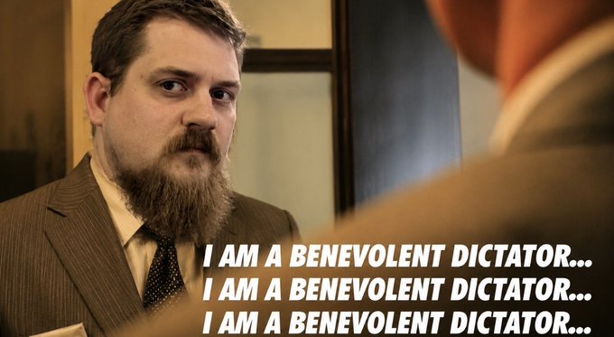 A bearded man in a suit looking at someone, with the caption 'I AM A BENEVOLENT DICTATOR...' repeated three times, appearing to be a still from a film or TV show.