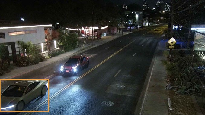 A nighttime traffic camera view showing a red car with bright headlights driving down a road, with an inset image of a silver sedan highlighted in an orange box.