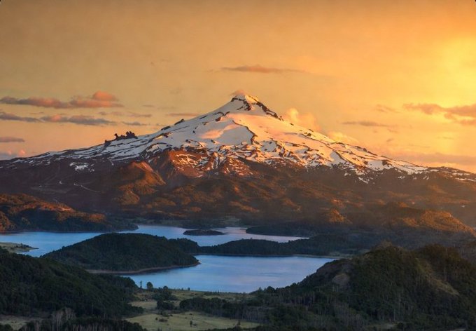 A scenic landscape photograph showing a snow-capped mountain illuminated by the golden light of sunset, overlooking a blue lake and forested valleys.