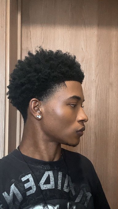 A profile view of a young man with curly dark hair, wearing a stud earring, a necklace, and a black t-shirt, standing against a wooden background.