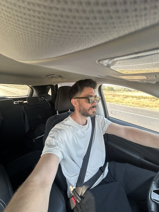 A bearded man wearing sunglasses and a white t-shirt takes a selfie while driving a car on a highway.