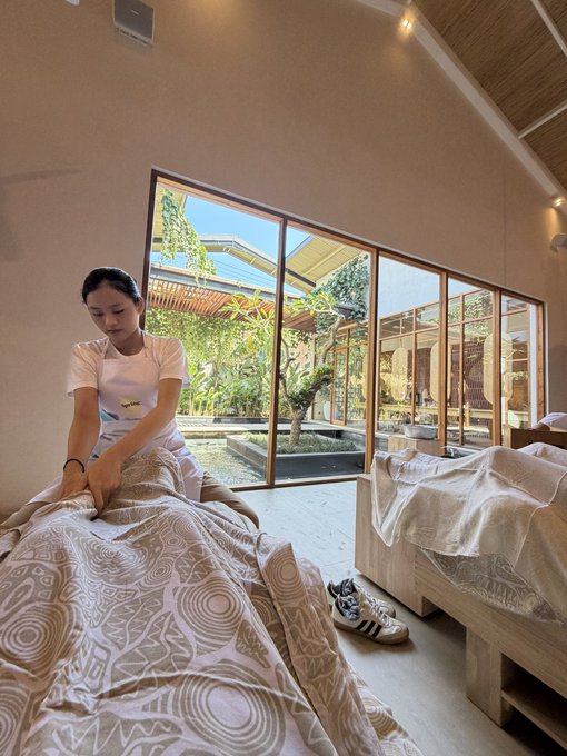 A woman in a white t-shirt gives a massage to a person lying on a bed inside a bright spa room with large windows looking out onto a garden.