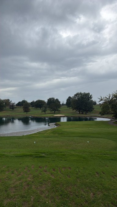 An outdoor photograph of a golf course featuring a small pond, green fairways, and trees under an overcast, cloudy sky.