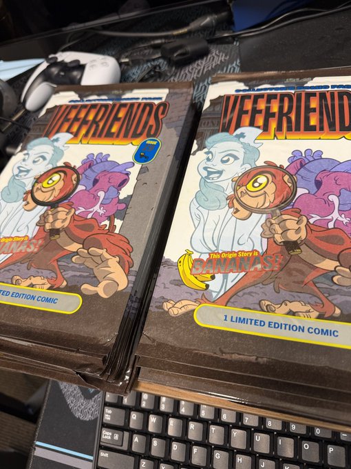 Stacks of 'WEEFRIENDS' limited edition comics featuring a banana-themed character, displayed on a desk near a keyboard and game controller.